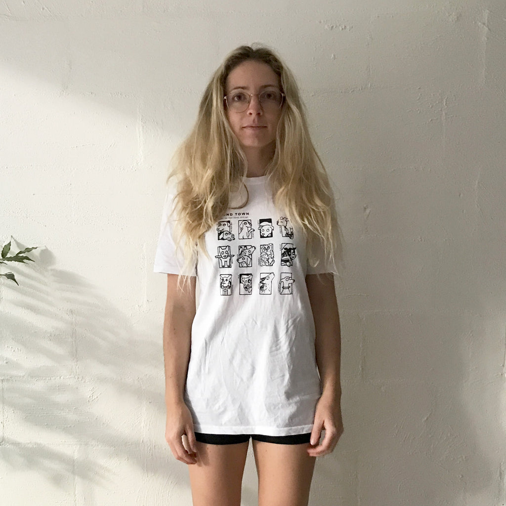 Blonde girl wearing a white tshirt with Hound Town black dogs cartoon artwork. She stands in front of a white brick wall and wears bike shorts and glasses.