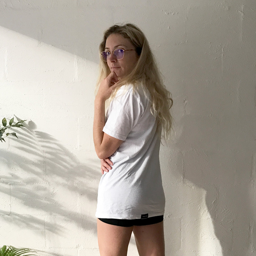 Hound Town tshirt being modelled by a blonde girl wearing glasses standing in front of a white brick wall with shadows. She is turning to look at the camera to show the back label on the tshirt.