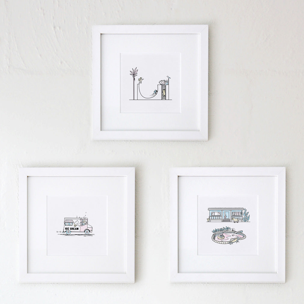 Three square white frames on a white wall holding fine art prints that feature cartoon illustrations by Clare Duffy including a halfpipe, Ice cream truck with music playing, a fishermans cottage with a pool in front, all scenes from Hound Town.